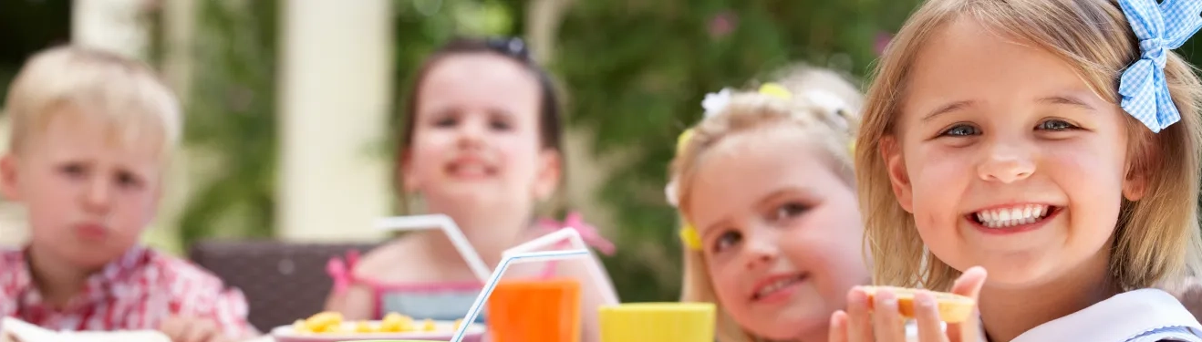 Lächelnde Kinder beim Essen im Freien, mit Getränken und Snacks auf einem Tisch.