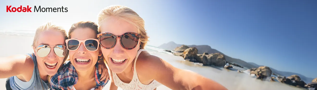 Drei lachende Frauen mit Sonnenbrillen genießen einen sonnigen Tag am Strand und machen ein fröhliches Selfie.