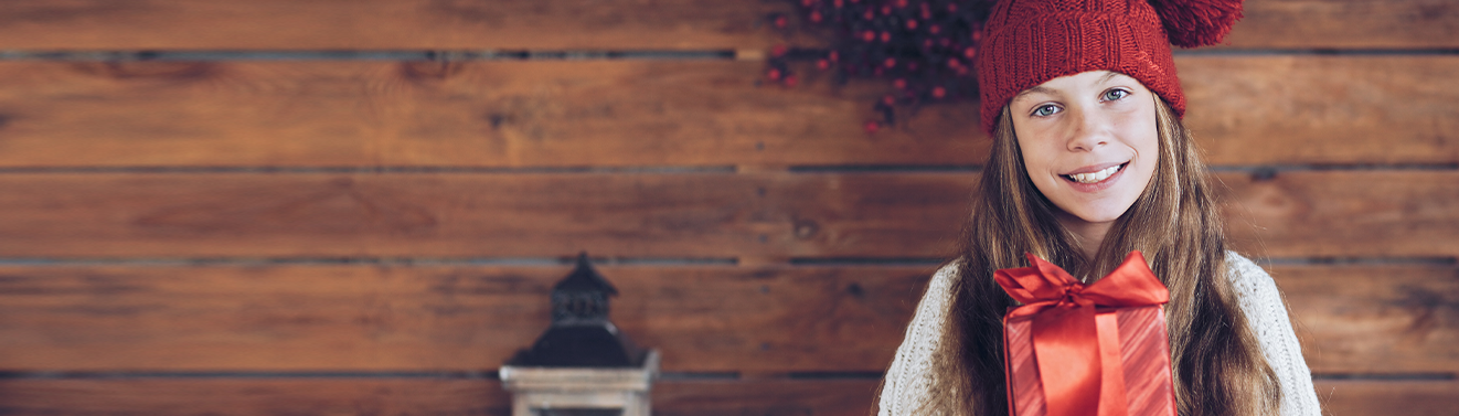 Kind mit roter Mütze hält ein verpacktes personalisiertes Weihnachtsgeschenk mit großer roter Schleife in den Händen.