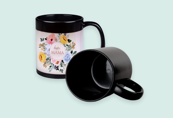 Magic Cup mit floralem Motiv, das erst beim Einfüllen heißer Getränke erscheint. Daneben liegt eine komplett schwarze Tasse.