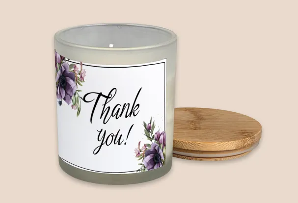 Duftkerze mit Deckel als Geschenkidee zur Hochzeit und Aufschrift ‚Thank you!‘, umgeben von lila Blumen auf weißem Etikett.