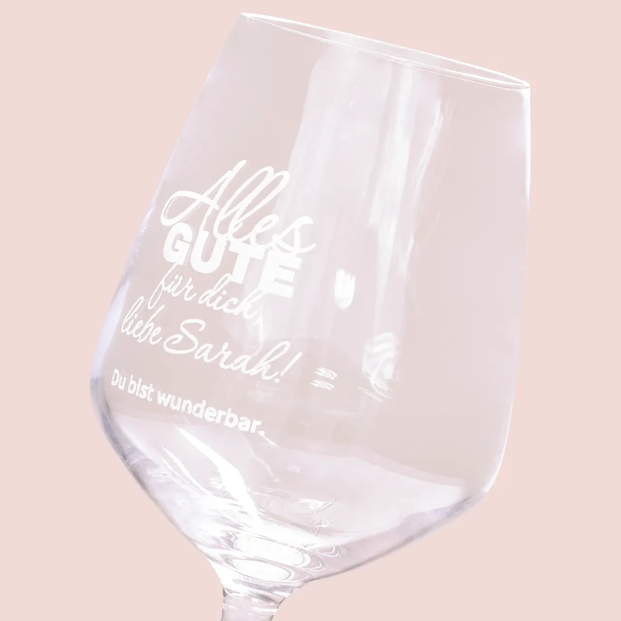 Nahaufnahme eines Weinglases mit dem Spruch ‚Alles Gute‘ auf Rosa-Hintergrund, ideal um Gläser personalisieren zu lassen.