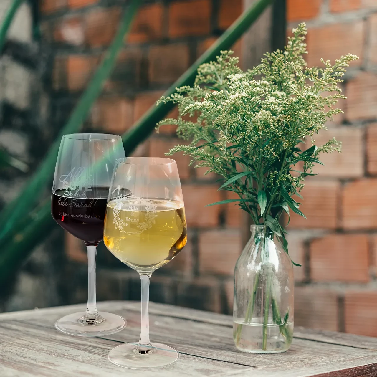 Zwei Rot- und Weißwein Gläser mit Gravur auf Holztisch neben einer Vase, individualisiert mit Namen und floralem Design.