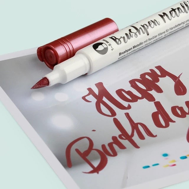Ein Brushpen in Weinrot-Metallic, mit dem „Happy Birthday“ auf eine Karte mit Hintergrunddekoration geschrieben wurde.