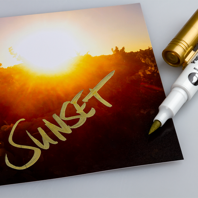 Ein Brushpen in Gold-Metallic, mit dem „Sunset“ auf ein Foto geschrieben wurde.